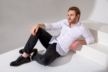 A man in a white shirt, dark jeans and black boots sits against a plain background and looks forward. Casual wear. Confident look. Psychology