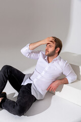 A man in a white shirt, dark jeans and black boots sits against a plain background, looks forward,...