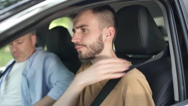 Concentrated young man fastening seat belt with blurred middle aged guy sitting on passenger seat in car. Portrait of confident Caucasian learner studying driving on sunny summer day outdoors.