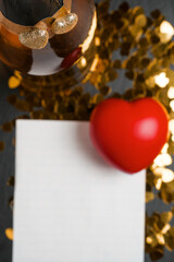 Two hearts with glitter on a bottle of champagne in focus. White paper, golden heart shaped confetti and read heart out of focus. Love and Saint Valentine background