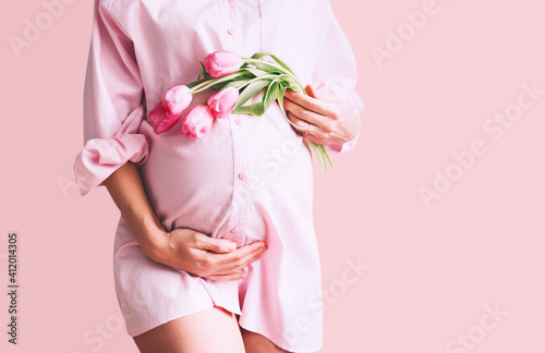 Pregnancy, Motherhood, Mother's Day Holiday concept. Young woman in maternity shirt dress with tulips flowers holds hands on belly. Beautiful pregnant woman waiting for baby birth.