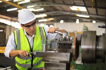 Asian mechanic worker in safety hard hat and reflective cloth is working with lathe machine inside the factory