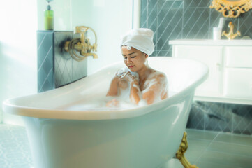 Naklejka premium Portrait beautiful young asian female massaging her hand with sponge in the tub. back front shot. Happy woman bathes in a bathtub white foam on her face fun emotions. Woman in bath and towel on head.