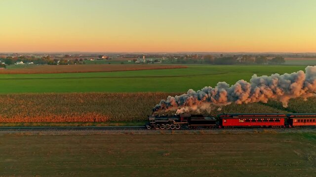 Aerial View of a Steam Engine with Passenger Cars at the Golden Sunrise Approaching with a Full Head of Steam and Smoke Traveling Thru the Farmlands Scaring a Herd of Cows
