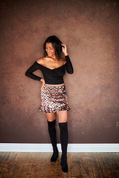 Stunning young African-American woman poses against warm toned wall wearing black off the shoulder blouse, bright skirt, and knee high boots - fashion - smiling