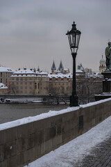 Fototapeta na wymiar view of snow-covered statues and street lights on the old stones of Charles Bridge on the Vltava River and in the background the snow-covered roofs of buildings and churches