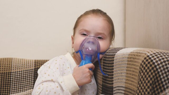 Treatment of a child at home for the flu and colds of the lungs. Kid will be inhaled with a nebulizer while sitting on the sofa in the children's room. A little girl breathes into a mask with medicine