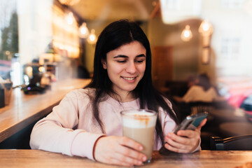 Girl smiling, drinks coffee in cafe and reading phone. Blurred background