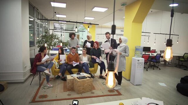 Professional startup team colleagues at meeting holding a paper documents having relaxed break on sofa in modern coworking space. Group looking at projects discussing building plan design project.