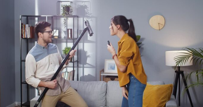 Portrait of Caucasian young happy married couple having fun in living room playing vacuum cleaner like guitar, dancing and singing while cleaning apartment, family concept, housekeeping