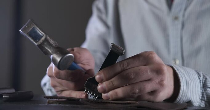 Man hits with Hammer. Makes Holes in Piece of Leather. Manufacturing of Leather Products. Manual Labor. Ancient Art. Work with Hands. Smooth Row of Thread Holes. Nice Seam. Precise Strikes. Master