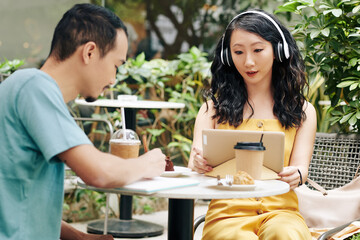 Pretty young Chinese woman with tablet computer making videocall when her boyfriend taking notes in textbook