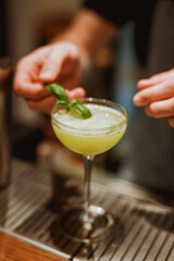 Bartender garnishing sour cocktail in a coupe glass with a basil leaf. Lifestyle vertical image. Selective focus.