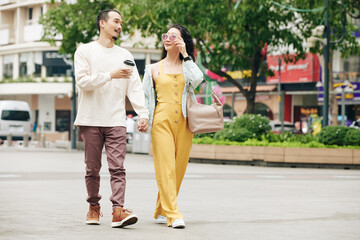 Stylish young Chinese couple holding hands and talking when walking in city on weekend