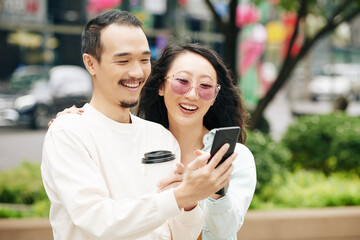Happy excited young Chinese couple with take out coffee taking selfie on smartphone when standing outdoors