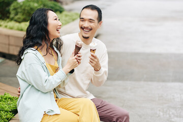 Laughing happy young Asian couple sitting on bench, talking and eating icecreams