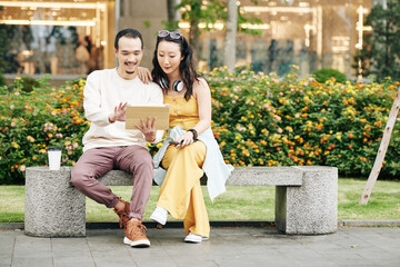 Fototapeta premium Cheerful young Chinese couple sitting on bench and discussing video or photos on tablet computer