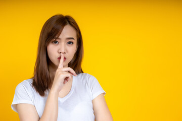 Young Asain woman requires silence. Young beautiful brunette has put forefinger to lips as sign of silence, isolated on yellow
