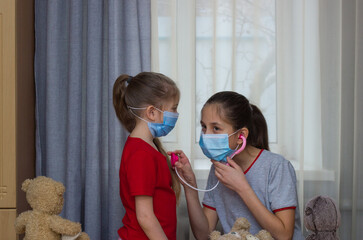 Two girls of school and preschool age, wearing medical masks, play in the hospital with toys, conduct a medical examination, listen in a phonendoscope.