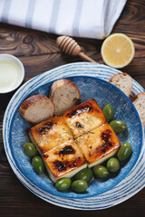 Blue bowl with baked feta cheese, green olives and baguette over brown rustic wooden background,...