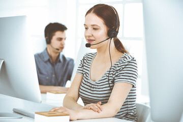 Fototapeta premium Casual dressed young woman using headset and computer while talking with customers online. Group of operators at work. Call center, business concept