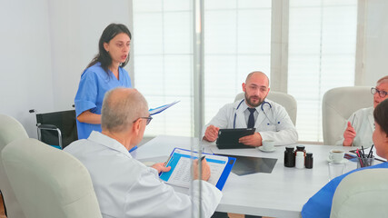 Woman medical assistant using clipboard in hospital conference room with coworkers explaining disease development. Clinic expert therapist talking with colleagues about disease, medicine professional