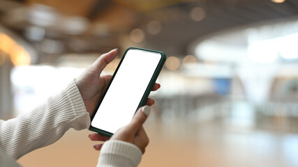 Close up view of young woman holding smart phone with empty screen on shopping mall blurred background.