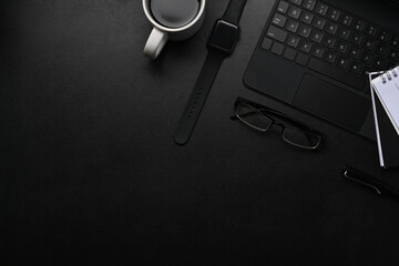 Above view of dark modern workspace with smart watch, notebook, wireless keyboard, glasses and copy space on black table.