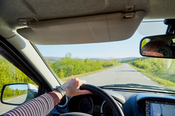 Car salon, steering wheel, hand of woman on it, mirror and view on landscape with road and nature at sunny summer or autumn day. Female traveller and driver during car trip. Concept of a single trip