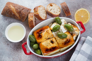 Blocks of baked feta cheese with giant green olives, olive oil and baguette, elevated view on a beige stone background