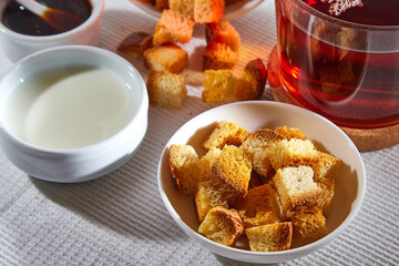 Square toasted pieces of homemade delicious rusk, hardtack, Dryasdust, zwieback, Liquid honey in a saucer, glass of milk and black tee in a cap on a white tablecloth.