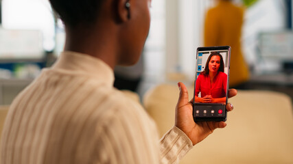 African woman explaining financial reports to remote manager on video call holding smartphone using headphones sitting on couch in business modern office. Diverse coworkers planning financial project