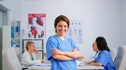 Portrait of young nurse standing in front of camera smiling in medical conference meeting office. Team of specialist doctors working in background talking about symptoms of disease in clinic room