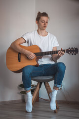 Young singer sits on a small stool in the corner and strums his song. Candid portrait of a blond man of European descent playing a guitar. Boredom during covid quarantine