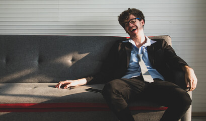 handsome caucasian businessman wearing black suit with loosen necktie sitting on a couch leaning against a backrest laughing happily. Success, employment concept
