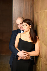 Fototapeta premium A couple in love. A bald man in a suit hugs a woman in a black dress against the wall. Valentine's Day celebration. Vertical photo