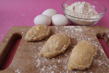 Raw cooking dumplings lie on a board beside egg flour on a pink background view from above