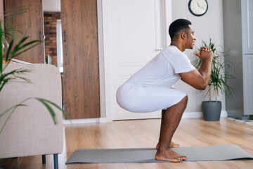 Side view of muscular muscular African-American man exercising and doing squats during working out at bright domestic room. Concept of sport training at home gym.