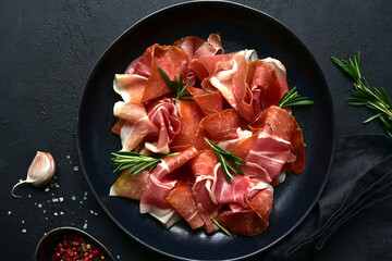 Slices of prosciutto di parma or jamon serrano (iberico)  on a black plate. Top view with copy...