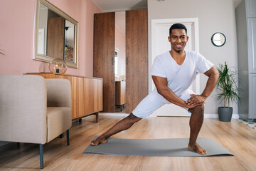 Front view of smiling strong African-American man doing side lunge exercise at home during working out standing on yoga mat at domestic room, looking at camera. Concept of sport training at home gym. - obrazy, fototapety, plakaty