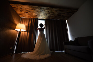 The bride in a wedding dress with her back turned, looking out the window. The interior is dark next to the lit lamp and she is wearing a white long wedding dress.