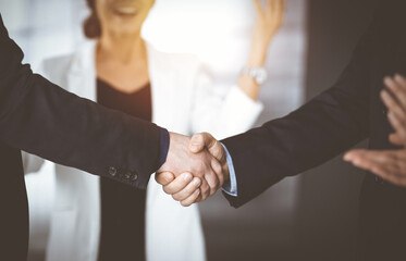 Unknown businesspeople are shaking their hands after signing a contract, while standing together in a sunny modern office, close-up. Business communication, handshake, and marketing concept