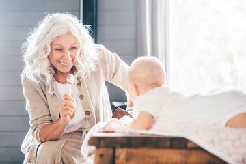 Grey haired grandmother playing with little baby girl on brown wooden table at home.