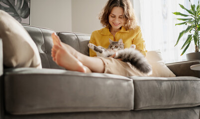 Beautiful cheerful young woman with a cute gray cat in her arms sitting on the couch at home, friendship and love for pets © olezzo