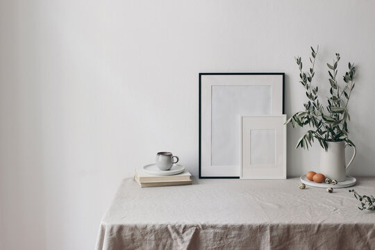 Spring, Easter breakfast still life. Cup of coffee, books and empty picture frames mockups. Linen tablecloth. Olive tree branches in ceramic jug. Hen, quail eggs. Farmhouse, Scandinavian interior.