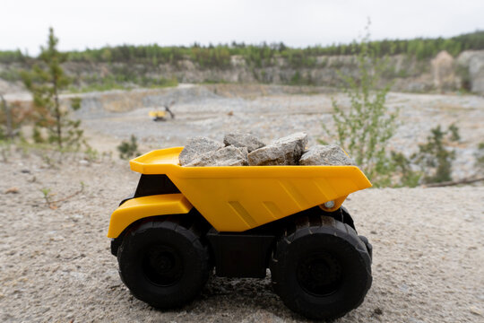 Closeup at small yellow car toy, mining truck with stones at the hill at the background of quarry. Stones and trees at background and blue sky.