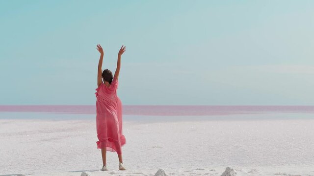 Beautiful paradise landscape of salt flats on pink lake with beautiful sky, looks like white desert. rear view Romantic dreamy woman in blowing pink dress with raised hands enjoying view and endless