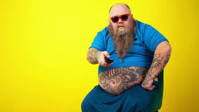 Funny man watching television. Big fat hipster with tattoo on yellow colored background