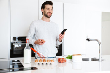 Happy young man using cellphone while cooking scrambled eggs