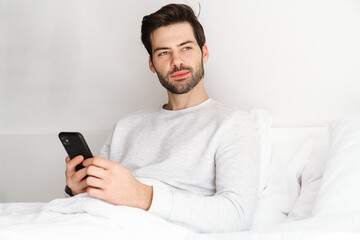 Pleased handsome man using mobile phone while resting in bed